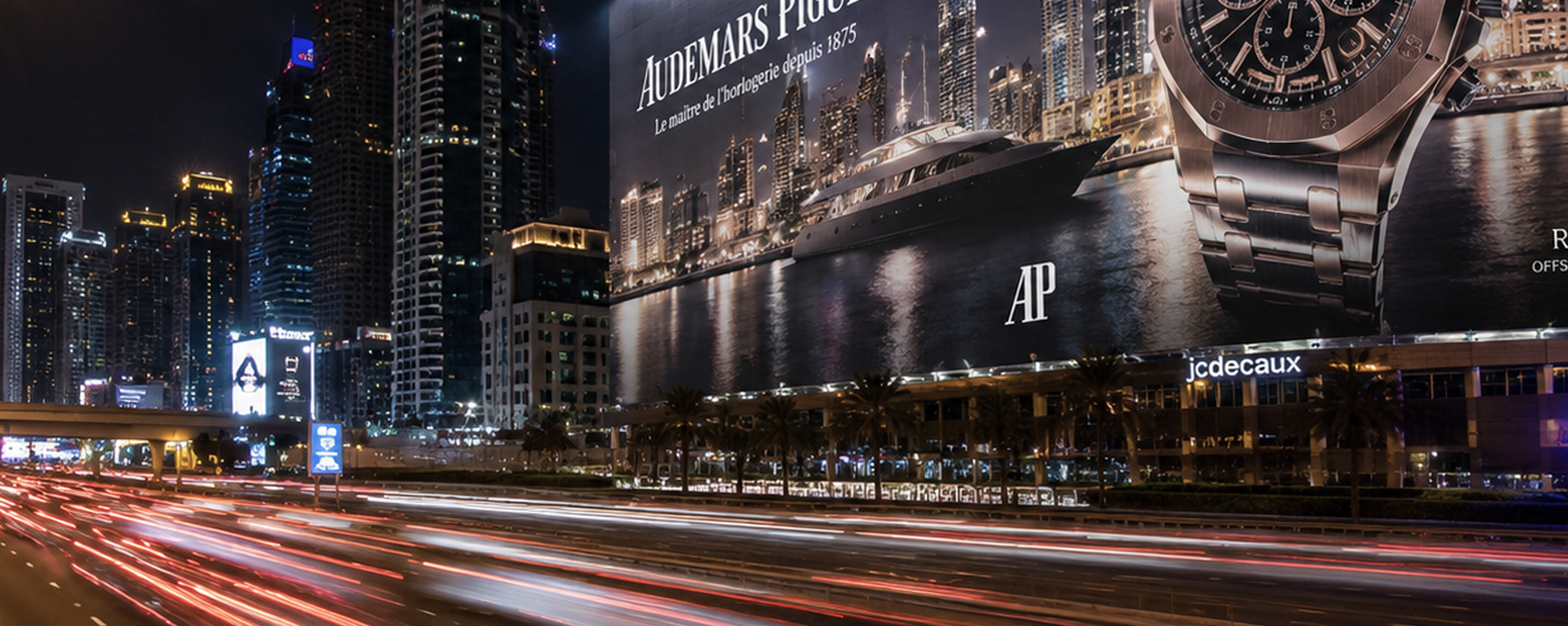 A luxury watch billboard towering over a night-time city marina skyline